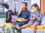 Little school girls sitting on bench in school yard and eating from lunch boxes.